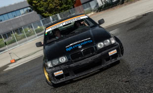 BMW nera in movimento su una strada bagnata, con edifici e alberi sullo sfondo.