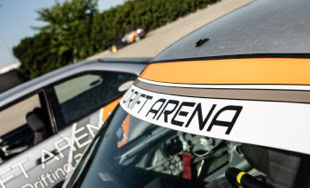 Due auto da corsa con adesivi "Drift Arena" sul parabrezza, parcheggiate su un piazzale asfaltato.