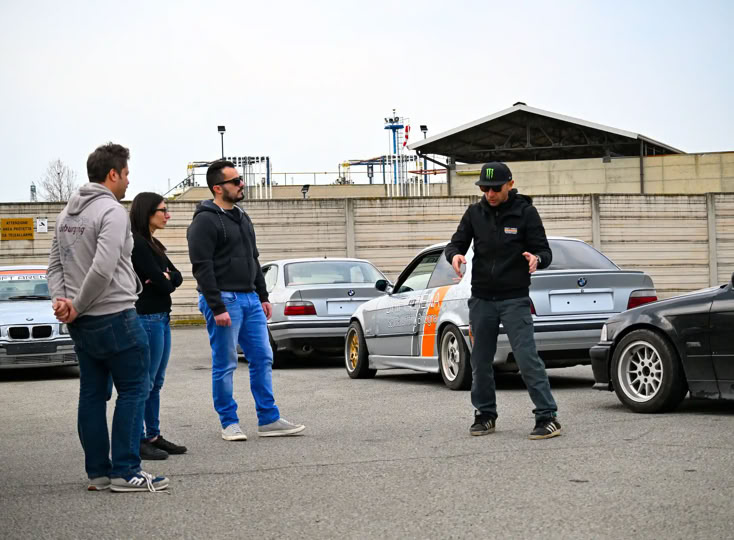 BMW Drifting nera con adesivi colorati parcheggiata su una strada, con un gruppo di persone in piedi vicino a un tendone bianco sullo sfondo.