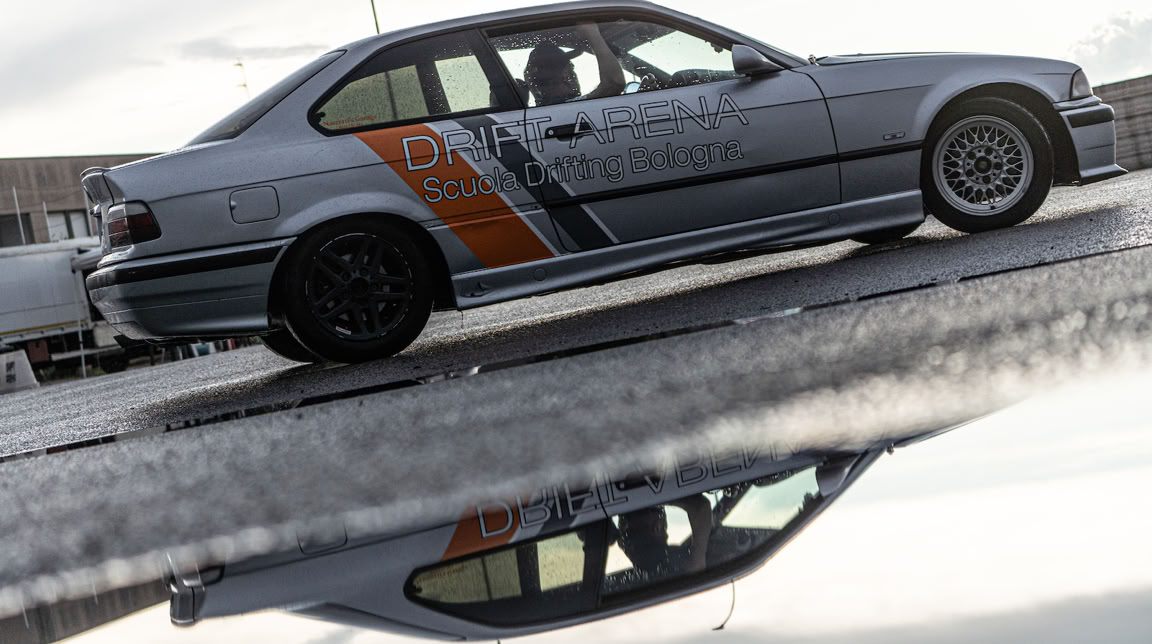 Auto sportiva grigia con adesivi arancioni e neri, riflessa su una superficie bagnata.