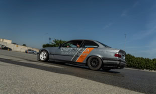 BMW e36 grigia da Drift con strisce arancioni e nere su una strada asfaltata, cielo blu sullo sfondo.