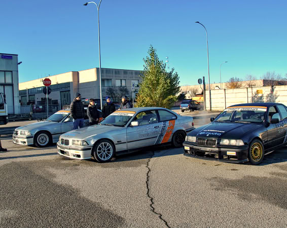 BMW Drifting nera con adesivi colorati parcheggiata su una strada, con un gruppo di persone in piedi vicino a un tendone bianco sullo sfondo.