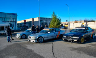 Tre auto sportive parcheggiate in un'area aperta con alcune persone sullo sfondo e un edificio industriale.