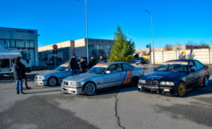 Tre auto sportive parcheggiate in un'area aperta con alcune persone sullo sfondo e un edificio industriale.