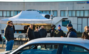 Un gruppo di persone in piedi all'aperto davanti a un furgone bianco con un tendone, con un'auto parcheggiata in primo piano.